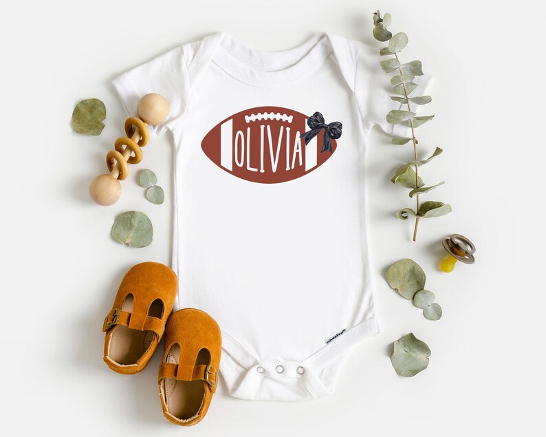 Puede incluir: Body de beb&eacute; blanco con un dise&ntilde;o de bal&oacute;n de f&uacute;tbol americano marr&oacute;n con el nombre "Olivia". Un lazo negro est&aacute; adjunto al costado. La imagen incluye zapatos de beb&eacute;, un juguete de madera y ramitas verdes.