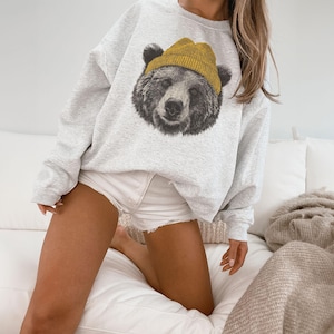May include: A light gray sweatshirt featuring a detailed graphic of a bear's face wearing a yellow beanie. The bear's fur is depicted in shades of gray and black. The sweatshirt is a casual style.