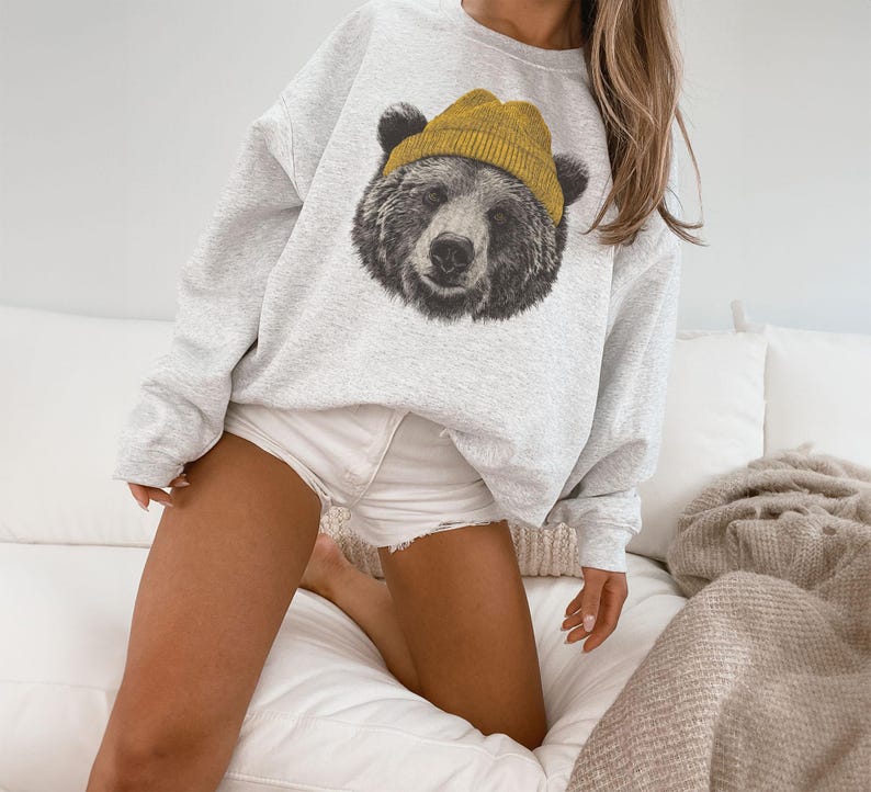 May include: A light gray sweatshirt featuring a detailed graphic of a bear's face wearing a yellow beanie. The bear's fur is depicted in shades of gray and black. The sweatshirt is a casual style.