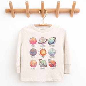 May include: A long-sleeved, cream-colored shirt featuring a colorful solar system design. The graphic includes illustrations of the sun and planets, each labeled with their names. The shirt is hanging on a wooden coat rack.