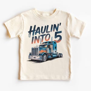 May include: Cream-colored t-shirt featuring a blue and orange semi-truck graphic. The text "HAULIN' INTO 5" is printed above the truck. The shirt is designed for children.