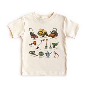 May include: A cream-colored short-sleeve t-shirt featuring a graphic print of various gardening and lawn care tools. The illustration includes a push mower, riding mower, zero-turn mower, leaf blower, string trimmer, shovel, rake, wheelbarrow, hedge trimmer, garden hose, watering can, and pruning shears, each labeled with its name.