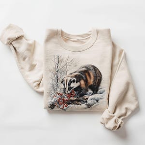 May include: Cream-colored sweatshirt featuring a detailed illustration of a badger eating red berries in a snowy landscape. The badger has black, white, and brown fur. The sleeves are rolled up. The sweatshirt is made of a soft material.
