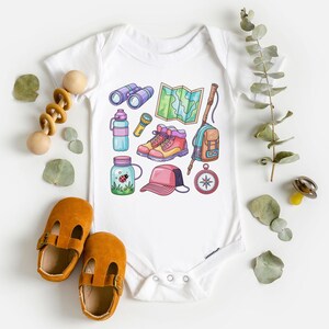 K&ouml;nnte beinhalten: Wei&szlig;er Baby-Body mit farbenfrohen Illustrationen von Wanderutensilien, darunter Fernglas, Karte, Stiefel, Rucksack und Kompass. Das Bild zeigt auch ein Paar braune Babyschuhe und ein Holzspielzeug.