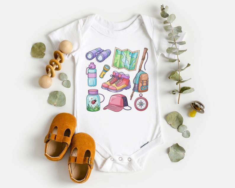 K&ouml;nnte beinhalten: Wei&szlig;er Baby-Body mit farbenfrohen Illustrationen von Wanderutensilien, darunter Fernglas, Karte, Stiefel, Rucksack und Kompass. Das Bild zeigt auch ein Paar braune Babyschuhe und ein Holzspielzeug.