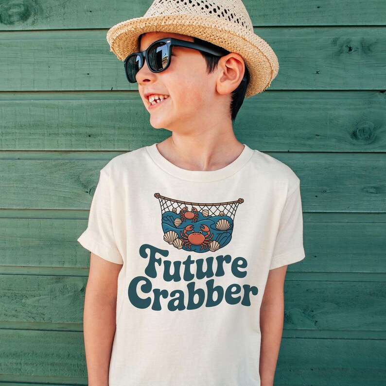 May include: A cream-colored t-shirt featuring a graphic of a crab in a net with seashells and the words "Future Crabber" in a retro font. The person in the image is wearing sunglasses and a straw hat.