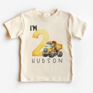 May include: A cream colored t-shirt with a yellow number two and a yellow dump truck. The text "I'm 2 Hudson" is printed on the shirt.