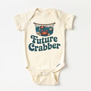 May include: Cream-colored baby bodysuit with a graphic of crabs, shells, and a net. The text "Future Crabber" is printed in teal. The bodysuit has short sleeves and a snap closure at the bottom.