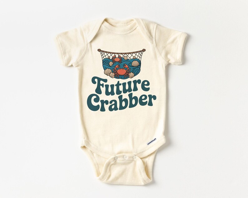May include: Cream-colored baby bodysuit with a graphic of crabs, shells, and a net. The text "Future Crabber" is printed in teal. The bodysuit has short sleeves and a snap closure at the bottom.