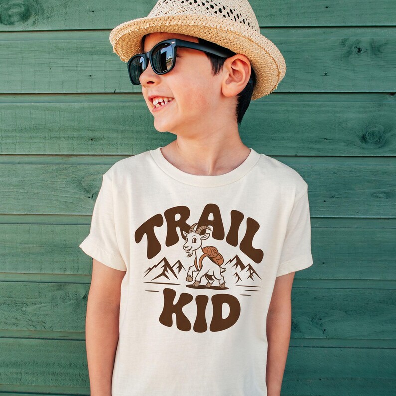 Peut inclure: Un t-shirt cr&egrave;me avec les mots "TRAIL KID" en marron, en texte arqu&eacute;. Une ch&egrave;vre de dessin anim&eacute; avec un sac &agrave; dos est centr&eacute;e sous le texte, avec des illustrations de montagnes. Le t-shirt est port&eacute; par un enfant portant un chapeau de paille et des lunettes de soleil.