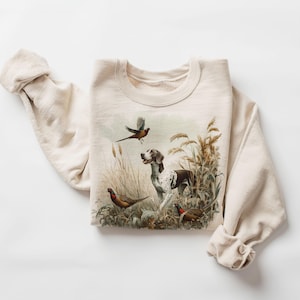 May include: A beige sweatshirt with a vintage illustration of a dog and birds in a field. The dog is brown and white with black spots. The birds are brown, white, and red.
