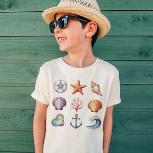May include: A cream-colored t-shirt featuring various sea-themed illustrations. The design includes a starfish, seashells, coral, an anchor, and a wave. The shirt is worn by a person wearing sunglasses and a straw hat.