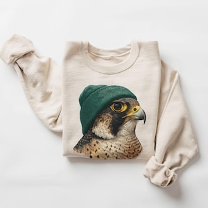 Könnte beinhalten: Cremefarbenes Sweatshirt mit einer detaillierten Illustration eines Falken mit grüner Mütze. Der Falke hat gelbe Augen und einen dunklen Schnabel. Die Ärmel sind hochgekrempelt. Das Sweatshirt liegt flach.