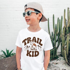 Peut inclure: T-shirt blanc avec un motif marron &laquo;&nbsp;TRAIL KID&nbsp;&raquo;. Le motif repr&eacute;sente une ch&egrave;vre avec un sac &agrave; dos devant des montagnes. Le t-shirt est port&eacute; par un enfant portant des lunettes de soleil et une casquette.