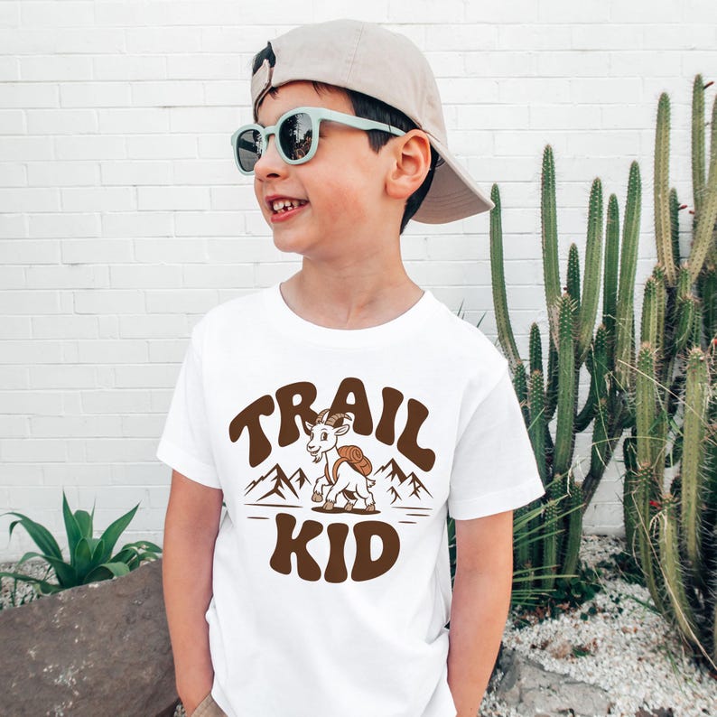 Peut inclure: T-shirt blanc avec un motif marron &laquo;&nbsp;TRAIL KID&nbsp;&raquo;. Le motif repr&eacute;sente une ch&egrave;vre avec un sac &agrave; dos devant des montagnes. Le t-shirt est port&eacute; par un enfant portant des lunettes de soleil et une casquette.