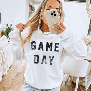 May include: White sweatshirt with the text "GAME DAY" in distressed black lettering. The long-sleeved top is a casual style, suitable for game day or everyday wear. The sweatshirt is a solid white color.