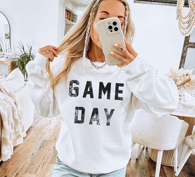 May include: White sweatshirt with the text "GAME DAY" in distressed black lettering. The long-sleeved top is a casual style, suitable for game day or everyday wear. The sweatshirt is a solid white color.