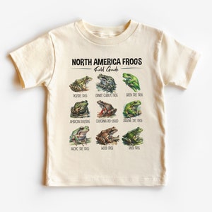 Könnte beinhalten: Ein beiges T-Shirt mit einem Grafikdruck eines Feldführers für nordamerikanische Frösche. Die Grafik zeigt Illustrationen von neun verschiedenen Fröschen mit ihren Namen unter jedem Bild. Die Namen sind: Pickerel-Frosch, Ornate Chorus-Frosch, Grüner Baumfrosch, Amerikanischer Ochsenfrosch, Kalifornischer Rotbeiniger Frosch, Grauer Baumfrosch, Pazifischer Baumfrosch, Wald-Frosch und Ochsenfrosch.