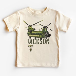 May include: A beige t-shirt with a green military helicopter and a parachuting figure. The text "JACKSON" is printed in green below the helicopter.