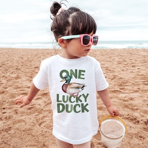 Puede incluir: Camiseta blanca con el texto "ONE LUCKY DUCK" en letras verdes. La camiseta presenta una ilustraci&oacute;n de acuarela de un pato real. El ni&ntilde;o lleva gafas de sol rosas y sostiene un cubo amarillo en una playa de arena.