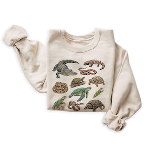 May include: A cream-coloured sweatshirt displaying illustrations of various reptiles. The design includes detailed drawings of an alligator, snakes, turtles, and lizards in different colours. The sweatshirt has a crew neck and long sleeves.