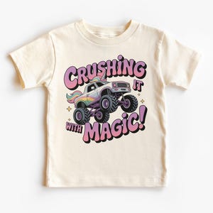 Puede incluir: Camiseta color crema con un gráfico de un camión monstruo con un cuerno de unicornio y detalles de arcoíris. El texto "Crushing it with Magic!" se muestra en una fuente de burbujas. La camiseta está diseñada para niños.