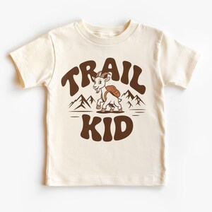 Peut inclure: T-shirt cr&egrave;me avec l'inscription "TRAIL KID" en marron, en forme d'arc. Un dessin anim&eacute; de ch&egrave;vre avec un sac &agrave; dos est en dessous du texte, avec des illustrations de montagnes en arri&egrave;re-plan. Le t-shirt est en mati&egrave;re souple.