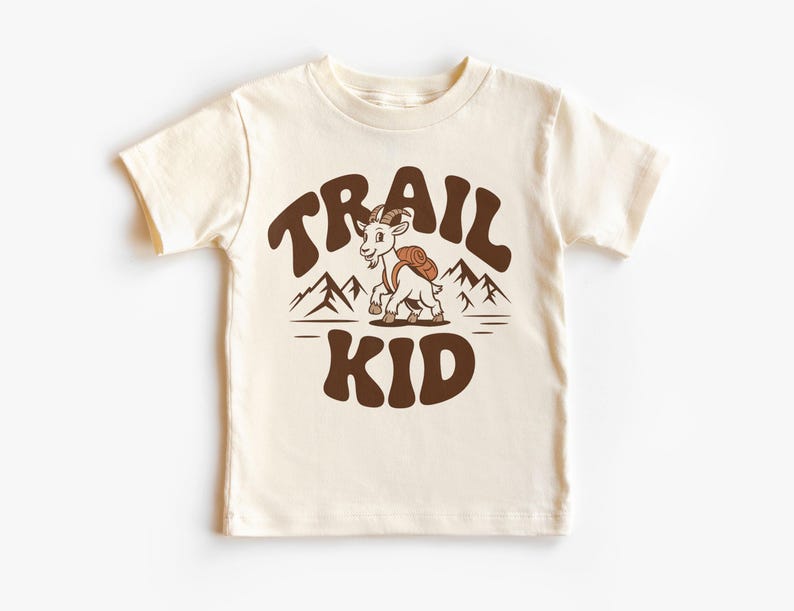 Peut inclure: T-shirt cr&egrave;me avec l'inscription "TRAIL KID" en marron, en forme d'arc. Un dessin anim&eacute; de ch&egrave;vre avec un sac &agrave; dos est en dessous du texte, avec des illustrations de montagnes en arri&egrave;re-plan. Le t-shirt est en mati&egrave;re souple.