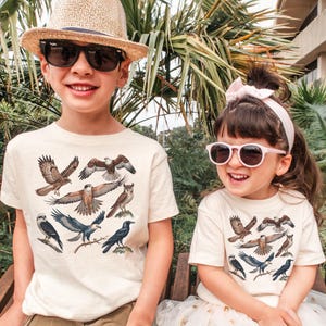K&ouml;nnte beinhalten: Zwei Kinder tragen cremefarbene T-Shirts mit einer detaillierten Illustration verschiedener Greifv&ouml;gel, darunter Falken, Eulen und Kr&auml;hen. Die Kinder tragen auch Sonnenbrillen und H&uuml;te. Der Hintergrund umfasst &uuml;ppiges gr&uuml;nes Laub.