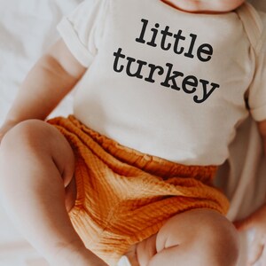 May include: A baby wearing a cream-colored onesie with the words "little turkey" printed in black. The baby is also wearing orange shorts. The baby is sitting on a white sheet.