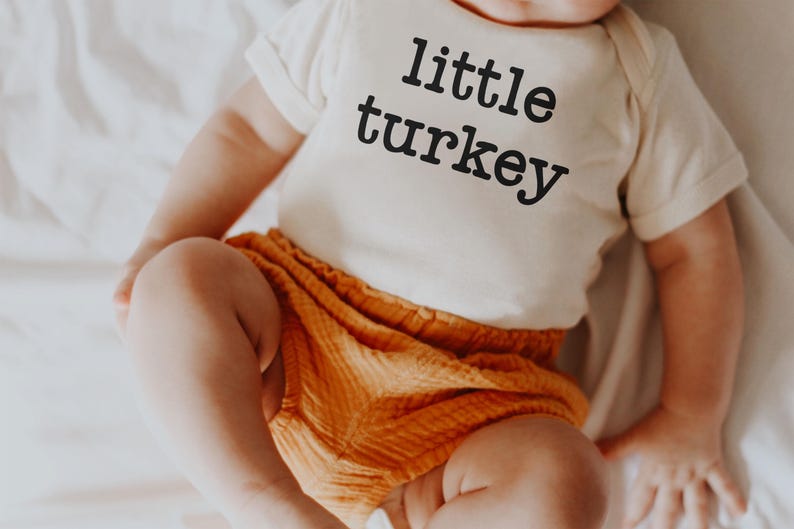 May include: A baby wearing a cream-colored onesie with the words "little turkey" printed in black. The baby is also wearing orange shorts. The baby is sitting on a white sheet.