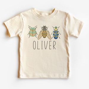 May include: A cream colored t-shirt with a bee and two beetles in watercolor style. The name "OLIVER" is printed below the insects.