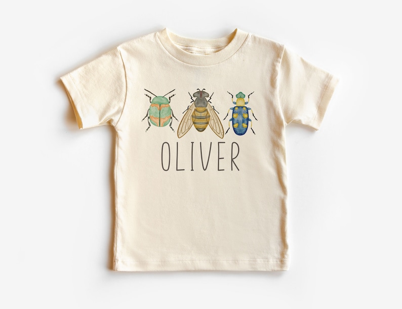 May include: A cream colored t-shirt with a bee and two beetles in watercolor style. The name "OLIVER" is printed below the insects.