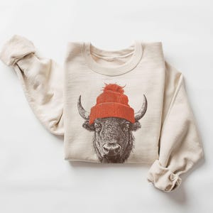 May include: Cream-colored sweatshirt featuring a detailed illustration of a bison wearing a bright orange knit cap with a pom-pom. The bison's face is the focal point, with its horns and facial features clearly visible. The sleeves are cuffed.
