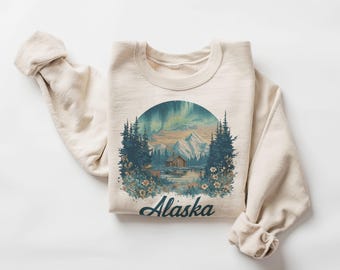 May include: A cream-coloured sweatshirt with a scenic illustration of Alaska. The design features a cabin, mountains, trees, and the word "Alaska" in a script font. The sleeves are rolled up, and the sweatshirt is displayed on a white background.