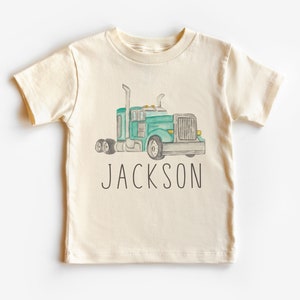 Puede incluir: Una camiseta de color crema con una ilustración de acuarela de un camión de plataforma verde y el nombre "JACKSON" impreso en negro.