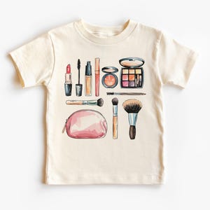 May include: A cream colored t-shirt with a colorful illustration of makeup brushes, lipstick, mascara, eyeshadow, and a makeup bag.