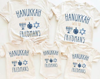 May include: Five cream-colored t-shirts and a onesie are displayed, each featuring the text "HANUKKAH WITH THE FRIEDMANS" in blue. The design includes a menorah, dreidel, and Star of David.
