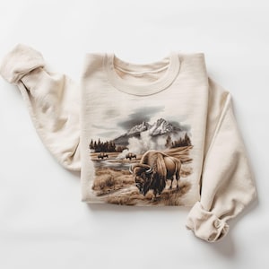 May include: A beige sweatshirt with a graphic print of a brown bison standing in a field with a mountain range in the background. The bison is facing the left side of the image. The mountain range is in the background and has a snowy peak. There is a steam rising from the ground in front of the bison.