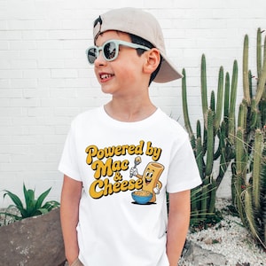 K&ouml;nnte beinhalten: Wei&szlig;es T-Shirt mit dem Text "Powered by Mac & Cheese" in Gelb und Orange. Die Grafik zeigt eine l&auml;chelnde Makkaroni-Nudel, die einen L&ouml;ffel &uuml;ber einer Sch&uuml;ssel Makkaroni mit K&auml;se h&auml;lt. Das T-Shirt wird von einem Kind getragen.