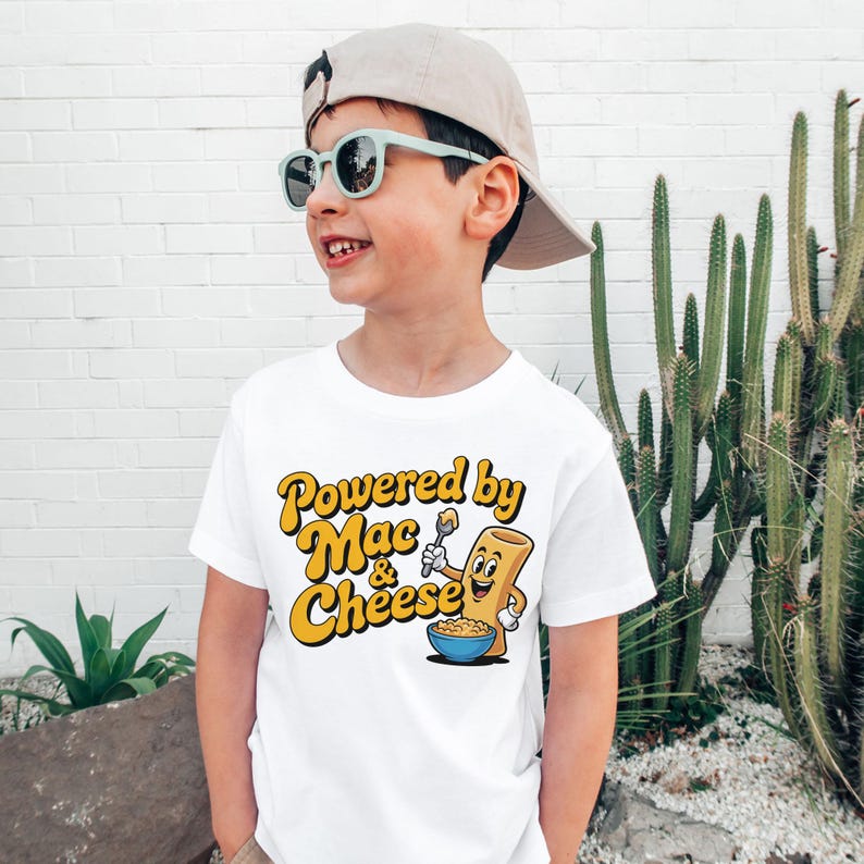 K&ouml;nnte beinhalten: Wei&szlig;es T-Shirt mit dem Text "Powered by Mac & Cheese" in Gelb und Orange. Die Grafik zeigt eine l&auml;chelnde Makkaroni-Nudel, die einen L&ouml;ffel &uuml;ber einer Sch&uuml;ssel Makkaroni mit K&auml;se h&auml;lt. Das T-Shirt wird von einem Kind getragen.