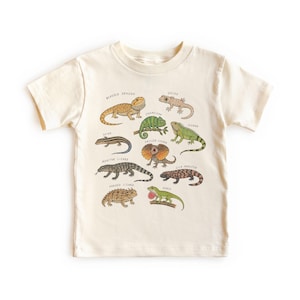 May include: A cream-colored short-sleeve t-shirt featuring a colorful graphic print of various lizards including a bearded dragon, gecko, chameleon, iguana, skink, frilled lizard, monitor lizard, gila monster, horned lizard, and an anole with labels.