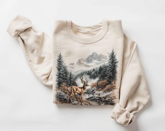 Puede incluir: Una sudadera beige con un estampado gráfico de un ciervo en un paisaje de montaña nevado. El ciervo es marrón con cuernos y está de pie en un bosque con árboles y nieve.