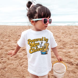 K&ouml;nnte beinhalten: Ein wei&szlig;es T-Shirt mit dem Text "Powered by Mac & Cheese" in Gelb und Orange. Das Shirt zeigt eine Cartoon-Makkaroni-Figur, die einen L&ouml;ffel &uuml;ber einer Sch&uuml;ssel Makkaroni mit K&auml;se h&auml;lt. Das Kind tr&auml;gt eine rosa Sonnenbrille.