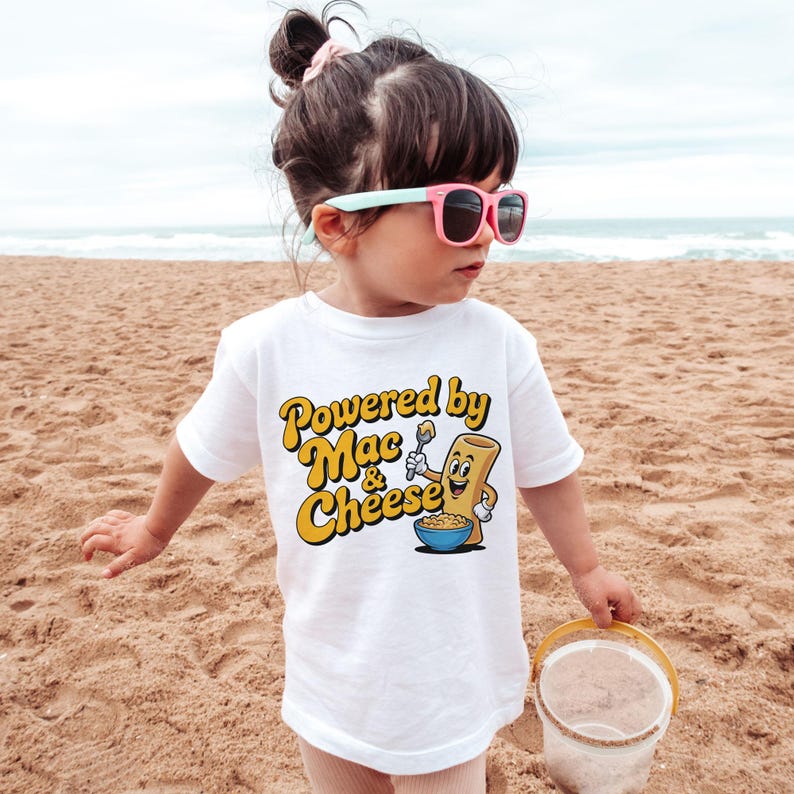 K&ouml;nnte beinhalten: Ein wei&szlig;es T-Shirt mit dem Text "Powered by Mac & Cheese" in Gelb und Orange. Das Shirt zeigt eine Cartoon-Makkaroni-Figur, die einen L&ouml;ffel &uuml;ber einer Sch&uuml;ssel Makkaroni mit K&auml;se h&auml;lt. Das Kind tr&auml;gt eine rosa Sonnenbrille.