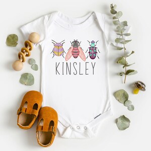 以下が含まれることがあります： 3匹のカラフルな虫のグラフィックと、その下に印刷された名前「Kinsley」が描かれた白いベビーロンパース。