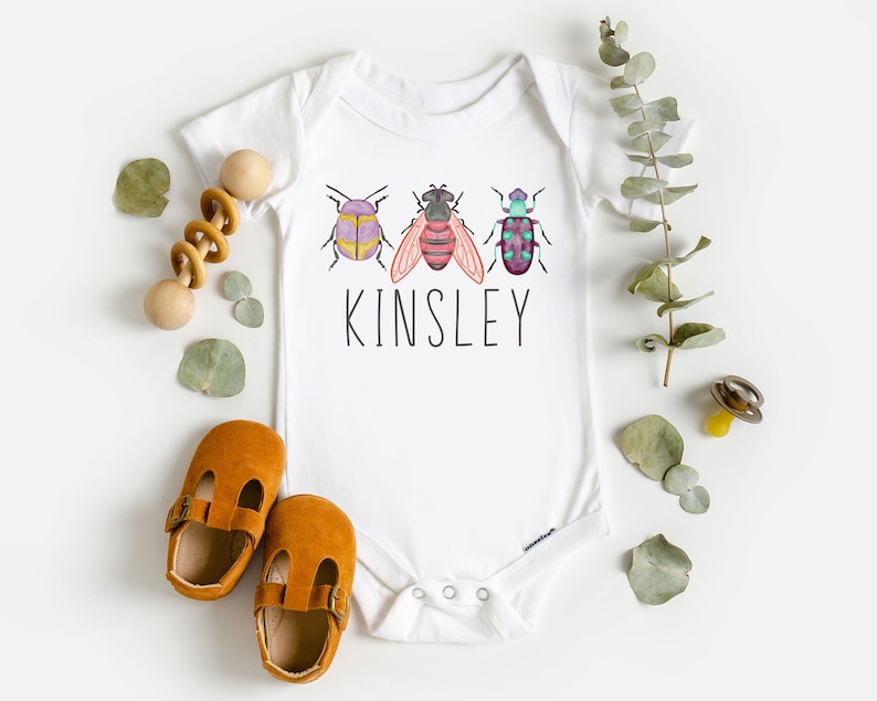 以下が含まれることがあります： 3匹のカラフルな虫のグラフィックと、その下に印刷された名前「Kinsley」が描かれた白いベビーロンパース。