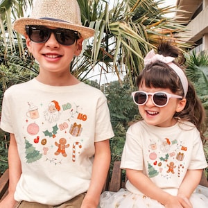 Puede incluir: Dos ni&ntilde;os con camisetas color crema con un gr&aacute;fico navide&ntilde;o. El gr&aacute;fico incluye a Pap&aacute; Noel, un hombre de jengibre, un &aacute;rbol de Navidad y la palabra "HO". Los ni&ntilde;os llevan gafas de sol y sonr&iacute;en.
