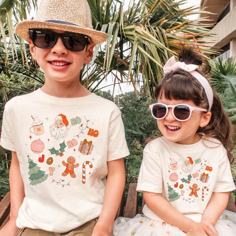 Puede incluir: Dos ni&ntilde;os con camisetas color crema con un gr&aacute;fico navide&ntilde;o. El gr&aacute;fico incluye a Pap&aacute; Noel, un hombre de jengibre, un &aacute;rbol de Navidad y la palabra "HO". Los ni&ntilde;os llevan gafas de sol y sonr&iacute;en.