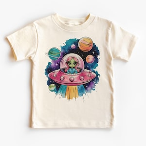 Könnte beinhalten: Ein weißes T-Shirt mit einem bunten Motiv eines grünen Alien-Mädchens, das in einem rosafarbenen Raumschiff durch eine Galaxie aus Sternen und Planeten fliegt.